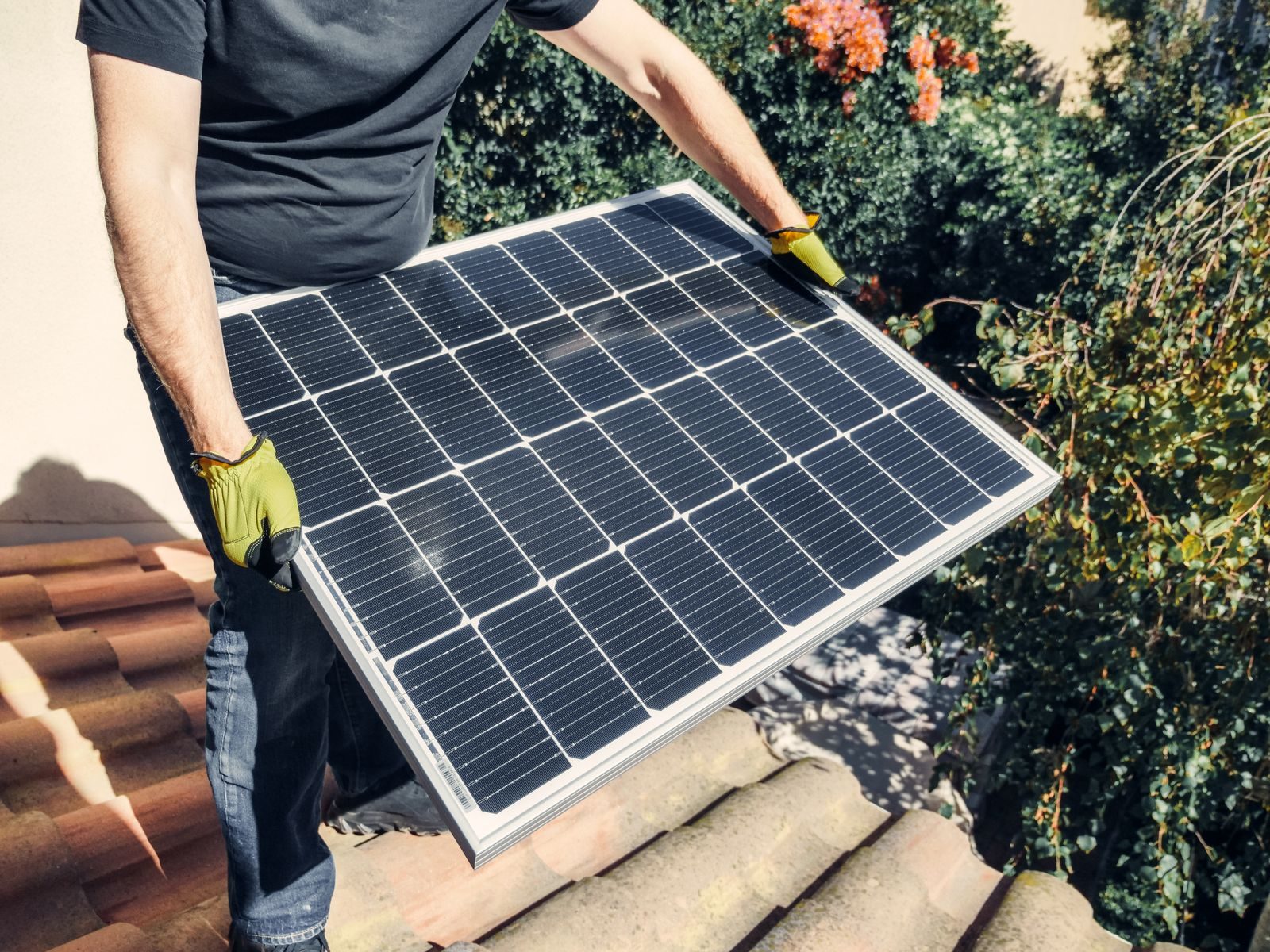 Solar installer in work gloves carrying a solar panel across a clay tile roof